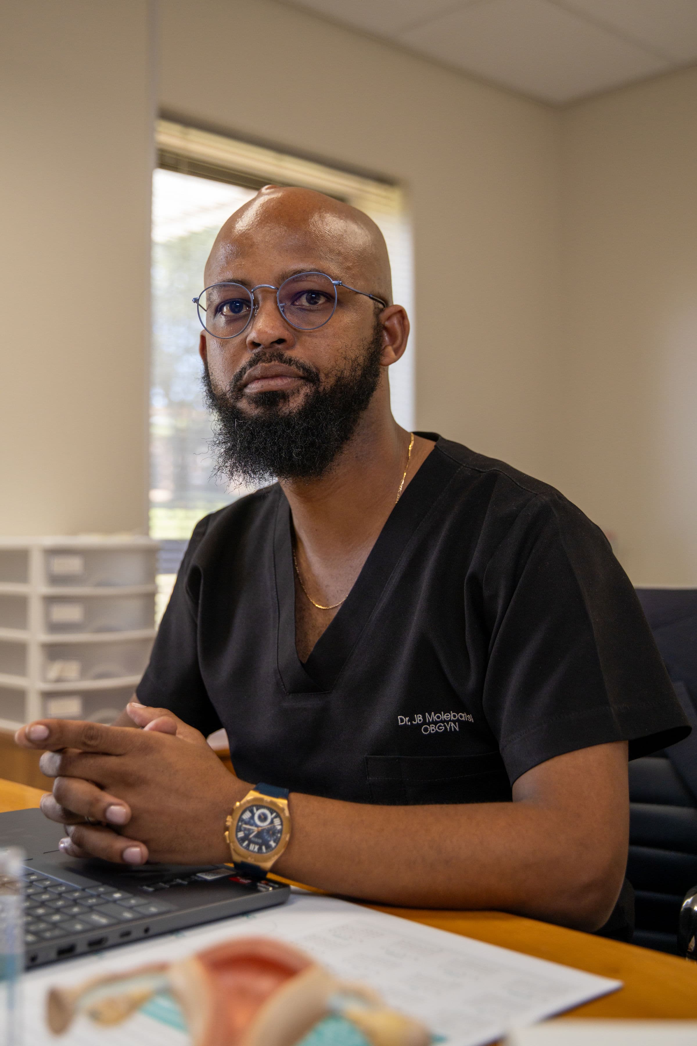 Dr. JB Molebatsi, a bearded OBGYN in black scrubs, sits at a desk.
